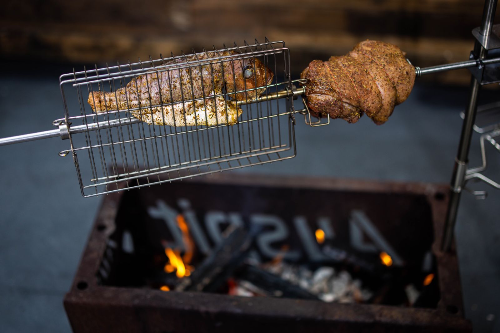 Auspit rotisserie basket holding a whole snapper over a fire pit, with a seasoned roast rotating beside it.
