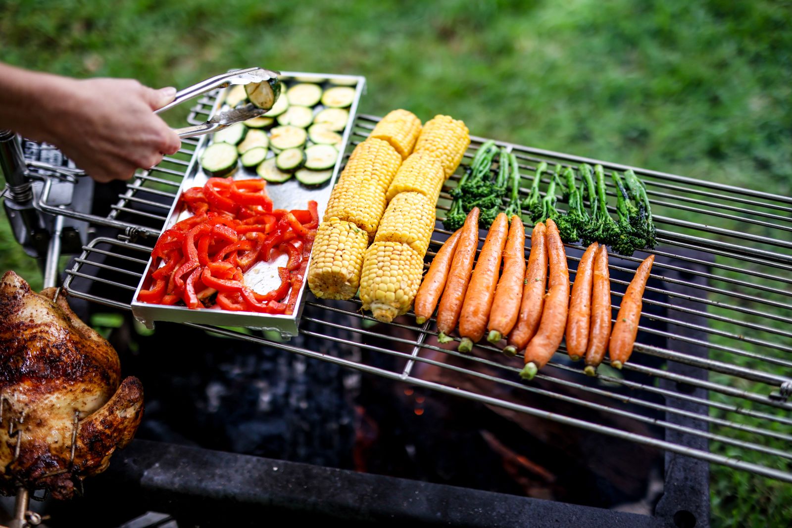 Grilled vegetables including carrots, corn, broccolini, zucchini, and capsicum cooking over an Auspit plate and grill combo, with rotisserie chicken visible on the side.