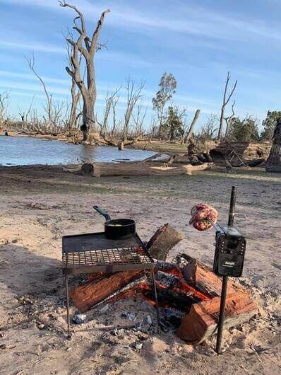 Auspit Spit Rotisserie cooking meat over open campfire with grill plate beside remote lakeside campsite