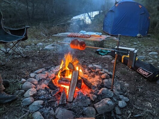 Portable Auspit spit rotisserie cooking meat over an open campfire, set up in a rocky fire pit near a river. Camping gear, chair, and tent visible in the background.