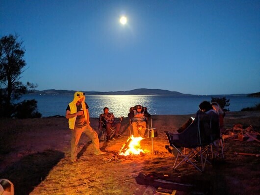 Group of campers relaxing around a campfire under the moonlight near a lake, enjoying the warmth of the fire with camping chairs and gear set up on the shore.