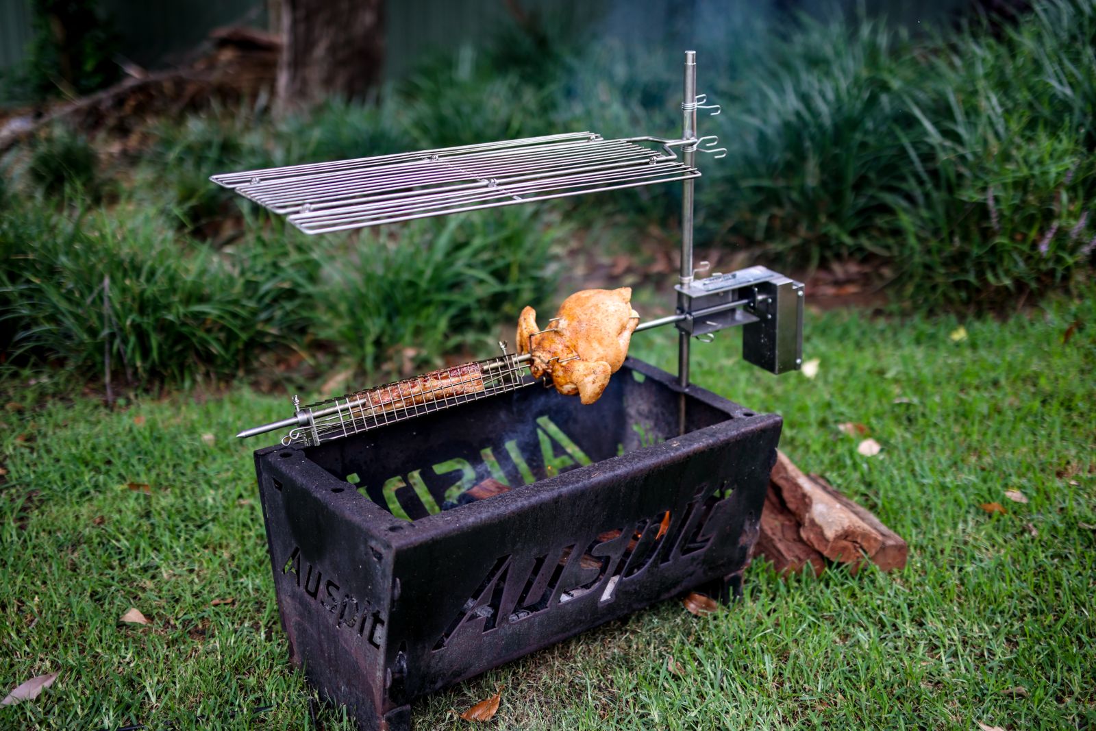 Auspit rotisserie set up with a whole chicken rotating over a fire, with a folding grill and stainless steel support post in the background.