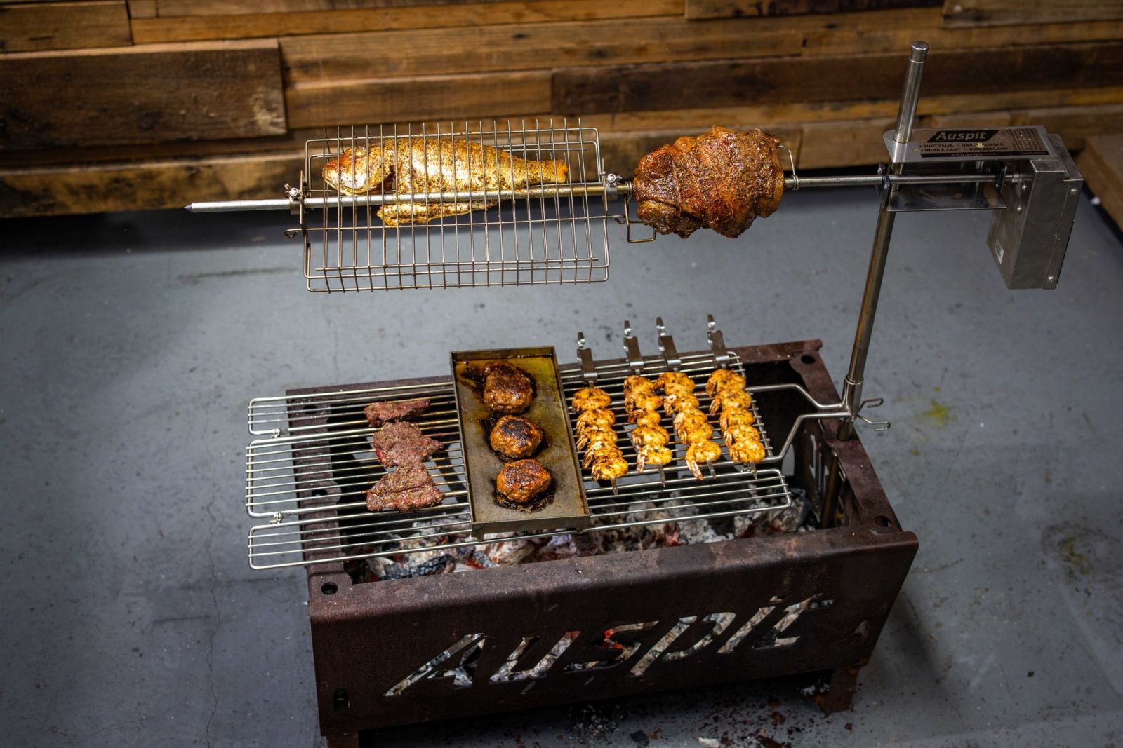 A complete Auspit rotisserie setup cooking a variety of food over charcoal, including a whole snapper, roast meat, prawns, burger patties, and steak pieces. The stainless steel rotisserie and grill show the versatility of the Auspit for outdoor cooking. A complete Auspit rotisserie setup cooking a variety of food over charcoal, including a whole snapper, roast meat, prawns, burger patties, and steak pieces. The stainless steel rotisserie and grill show the versatility of the Auspit for outdoor cooking.