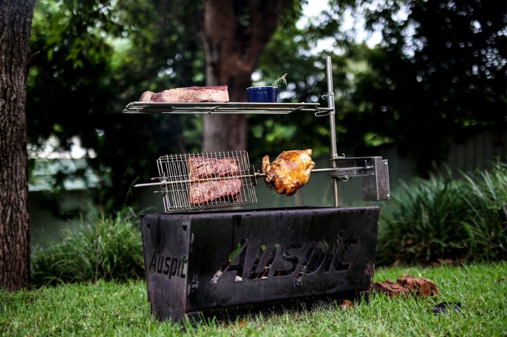 Auspit rotisserie setup in a grassy backyard, featuring a chicken and meat roasting over charcoal. A cooking basket holds additional meat, and a grill rack above displays a steak and a small pot with herbs.