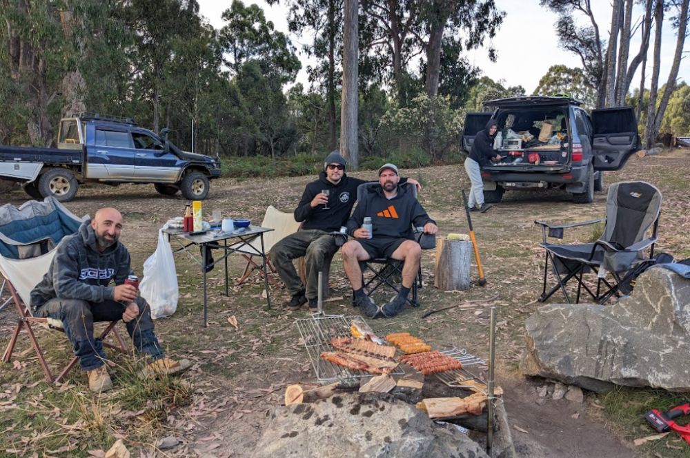 Group of men enjoying a camping trip in the bush, sitting around an Auspit rotisserie setup grilling sausages, ribs, and meat skewers over a campfire. The campsite is surrounded by trees with 4WD vehicles and camping gear in the background, creating a relaxed and rustic outdoor cooking scene.