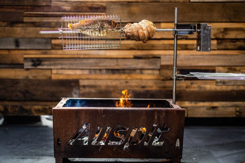 Auspit rotisserie cooking setup with fish secured in a basket and a seasoned rolled roast rotating over an open flame. The rust-colored fire pit features a cutout "Auspit" logo and stands in front of a wooden plank wall. A side tray is attached to the rotisserie post.