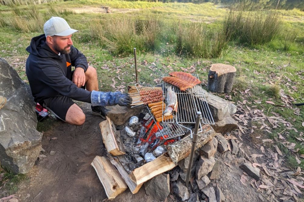 Man cooking with an Auspit rotisserie setup at a campsite, grilling sausages, fish, and ribs over an open fire. The rotisserie system is anchored with support posts and positioned over a fire pit built from rocks and logs. The scene is set in a grassy, wooded outdoor area.
