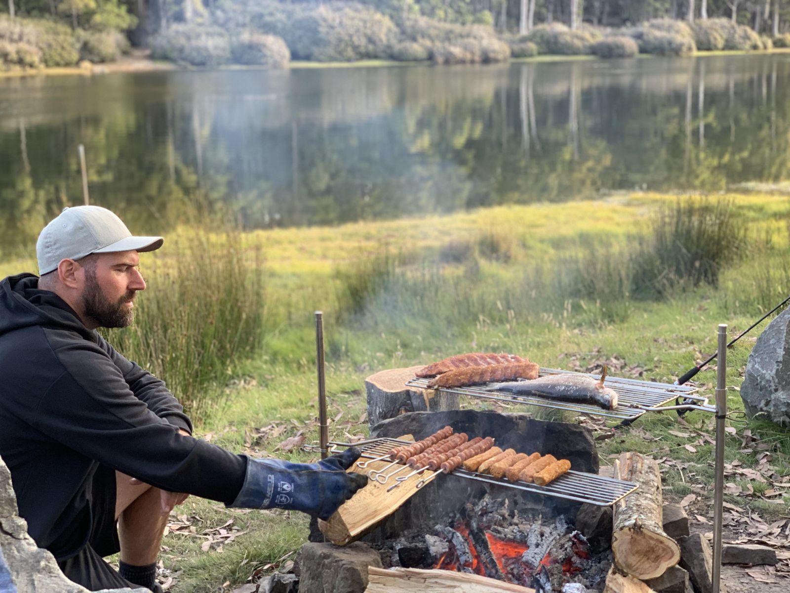 "Camping scene by a lake with a man cooking sausages and ribs on an Auspit rotisserie, using a stainless steel folding grill, while the other side holds fish. The man wears a cap and a black hoodie, kneeling next to the grill setup