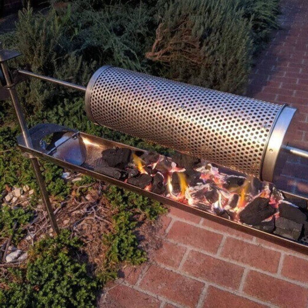 The image shows the Auspit Firetray with charcoal beneath the Auspit Spitmate Basket, set in an outdoor setting with a paved surface and greenery.