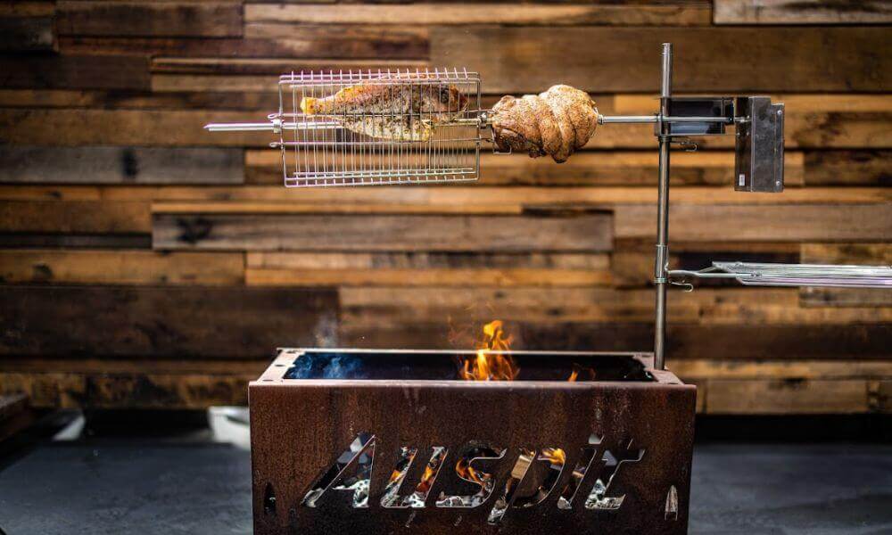 Auspit rotisserie kit featuring an accessory basket with meat on a spit, cooking over a fire in a rustic metal base.