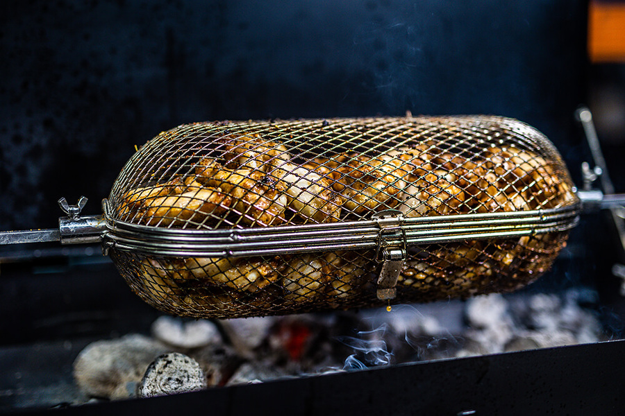 Auspit Cage Tumbler rotating over charcoal fire perfect for roasting vegetables, wings, and smaller cuts with even heat.