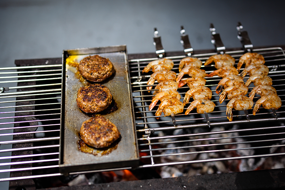 Seasoned prawns on skewers and burger patties grilling over charcoal on an Auspit setup.