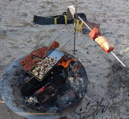 This is an image of the Auspit Camping spit cooking up 2 roasts, the hotplate filled with mushrooms and the swinging grill cooking steak over an open fire while travelling in Australia