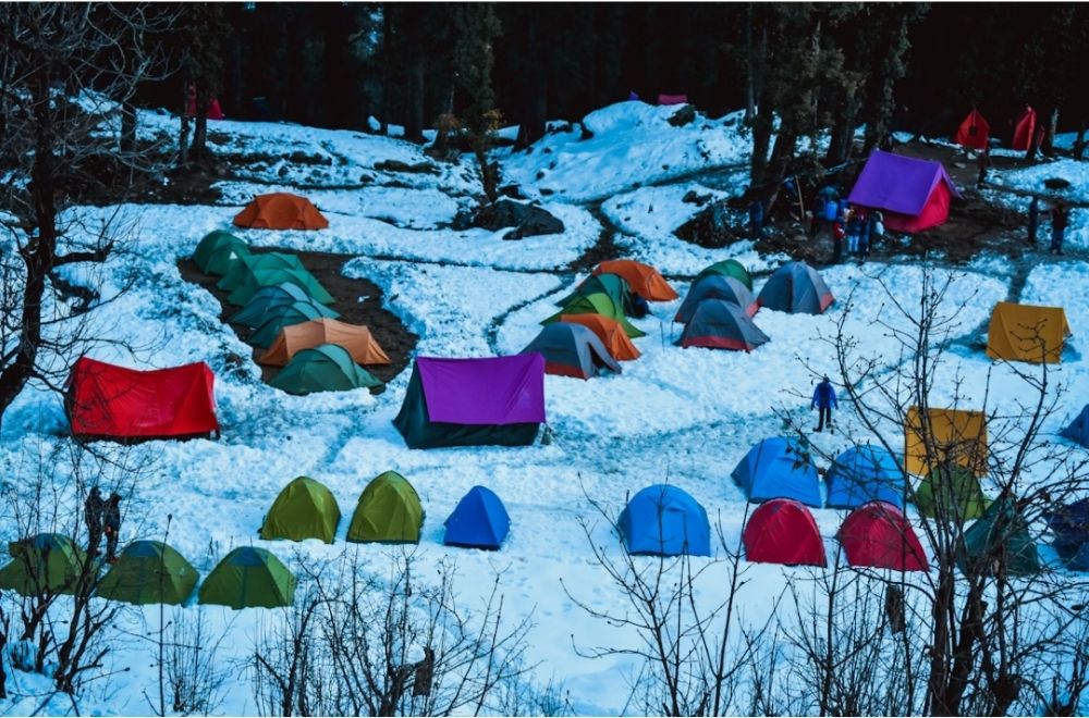 Colourful tents are scattered across a snowy landscape in a forested area, showing a busy winter camping scene with people walking around and gathering near tents.