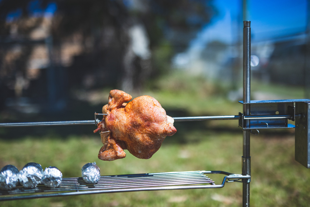 The image shows an Auspit Rotisserie with a chicken roasting on a spit over an open flame. The rotisserie includes a grilling rack with some foil-wrapped items, illustrating the versatility of the setup for outdoor cooking.