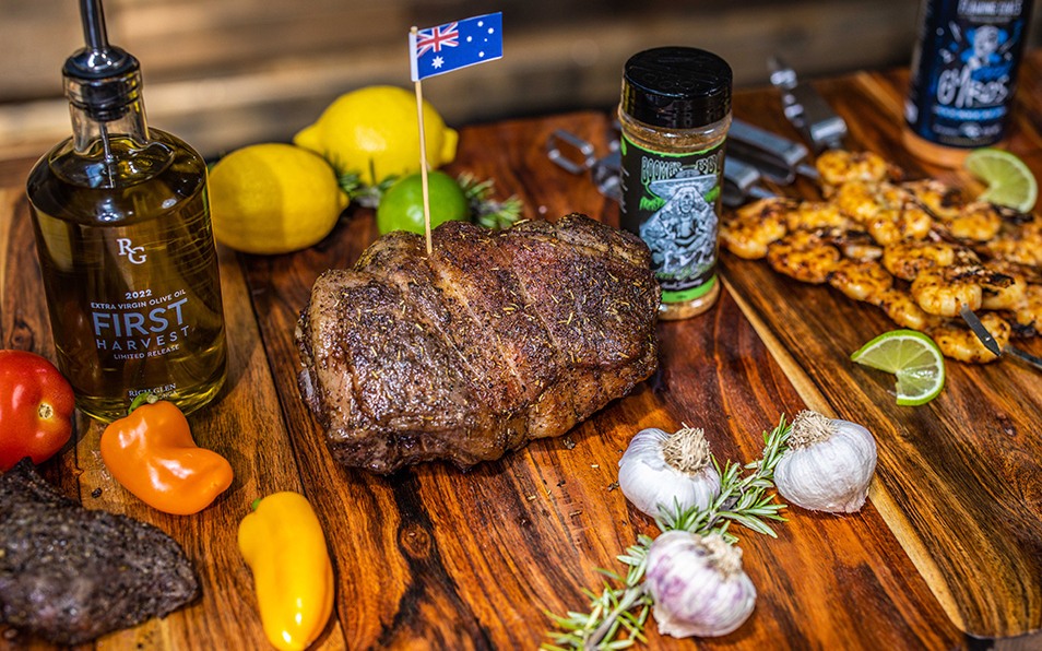 Perfectly cooked leg of lamb resting on a wooden board with garlic, rosemary, prawns, capsicum, olive oil, and Boomas BBQ seasoning beside it.