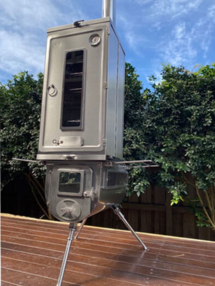Tall stainless steel Winnerwell wood stove with an attached vertical smoker box, standing on tripod legs on a wooden deck, with greenery and blue sky in the background.
