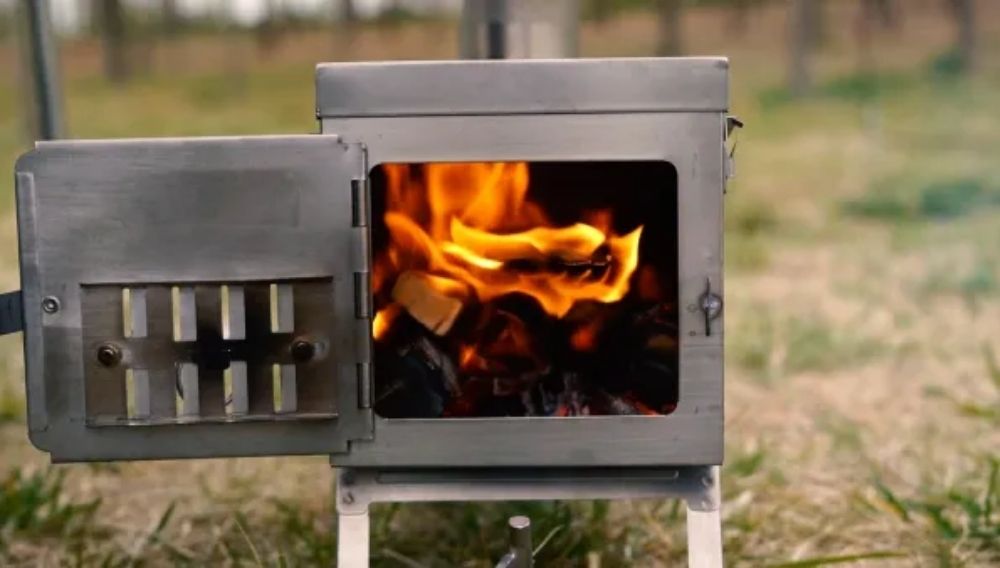 Fastfold Titanium Camping Stove by Winnerwell with its door open, showing a lively fire burning inside. The stove is set up outdoors on grassy ground with blurred trees in the background.