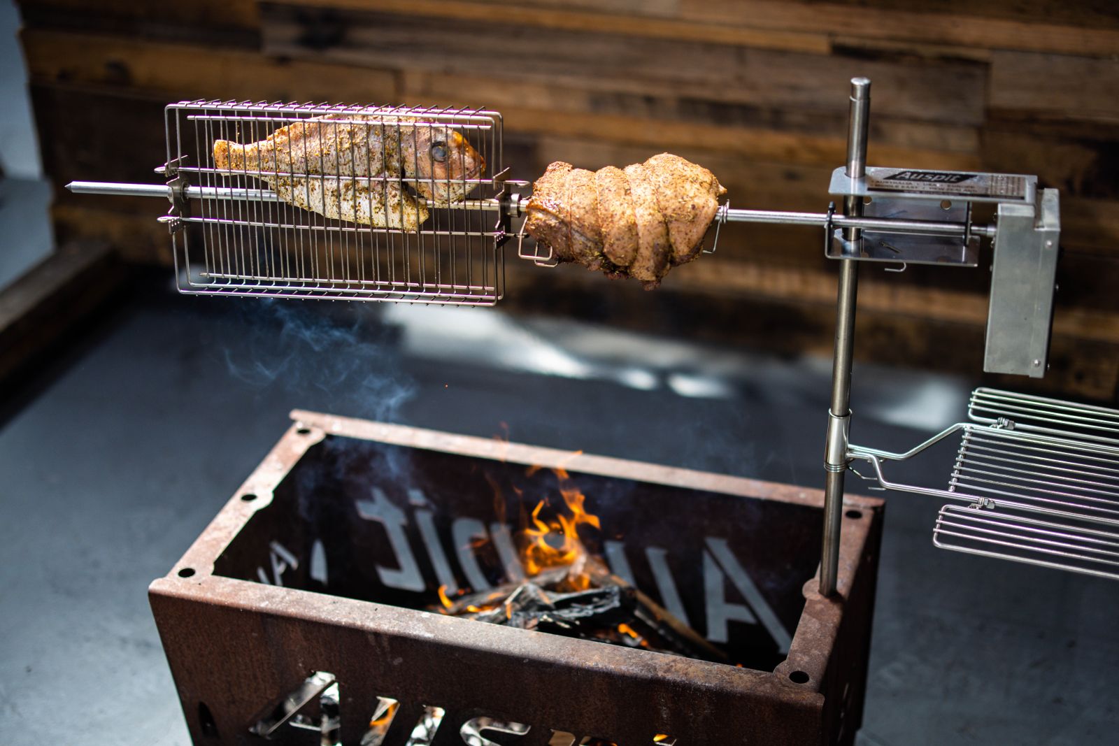 Auspit rotisserie with stainless steel motor cooking snapper and leg of lamb over an open fire in a steel fire pit.