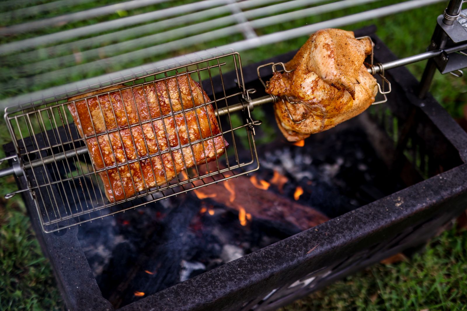 Rotisserie basket holding pork roast alongside a whole chicken on prongs, turning over a fire pit.