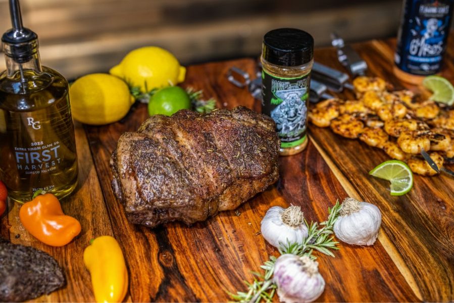 Roast lamb on a wooden board surrounded by Butcher’s Axe seasoning, olive oil, garlic, rosemary, and grilled prawn skewers.