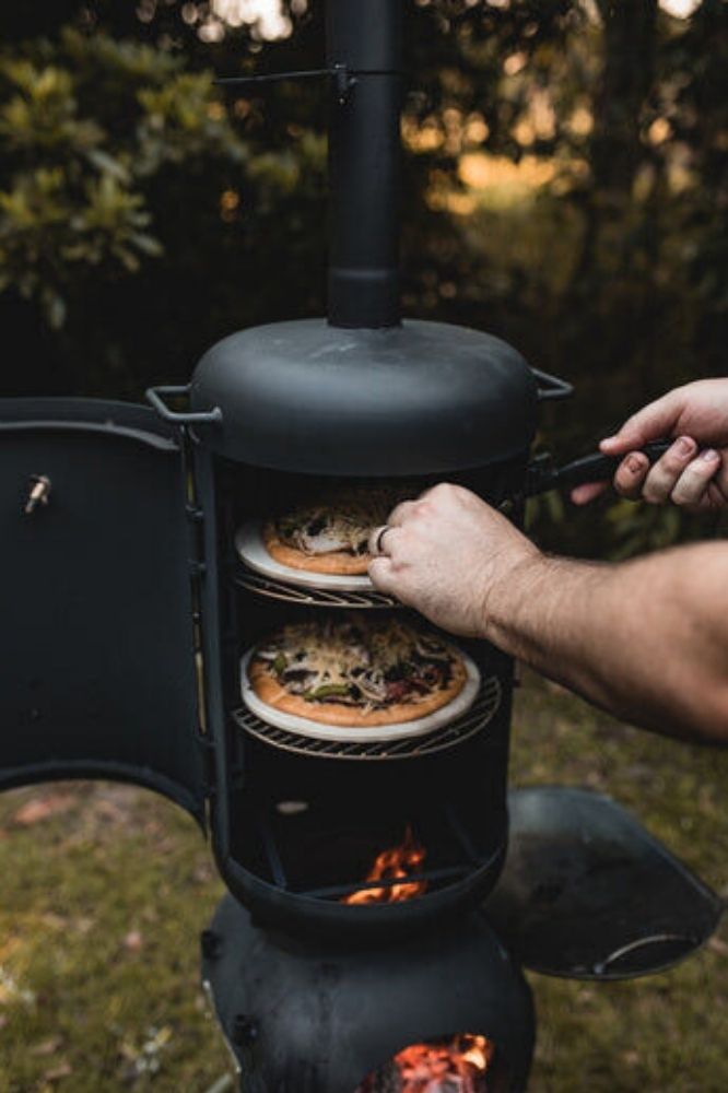 A person using a tool to rotate or remove a pizza from the top rack of an Ozpig Oven Smoker outdoors, with another pizza cooking below and visible flames in the firebox beneath.