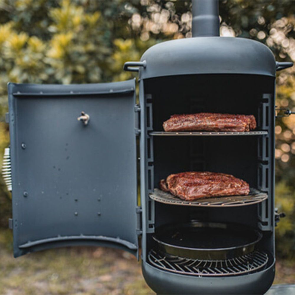 The_Ozpig_Oven_Smoker_with_its_door_open, showing_two_racks_of_seasoned_meat_smoking_inside_and_a_drip_tray_at_the_bottom.The_unit_is_set_outdoors_with_greenery_in_the_background.