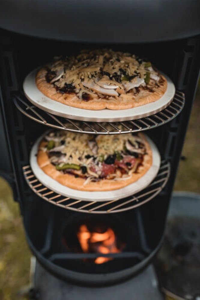 Two homemade pizzas topped with cheese, mushrooms, and vegetables are being cooked on separate racks inside the Ozpig Oven Smoker, with a visible flame burning below for wood-fired cooking.