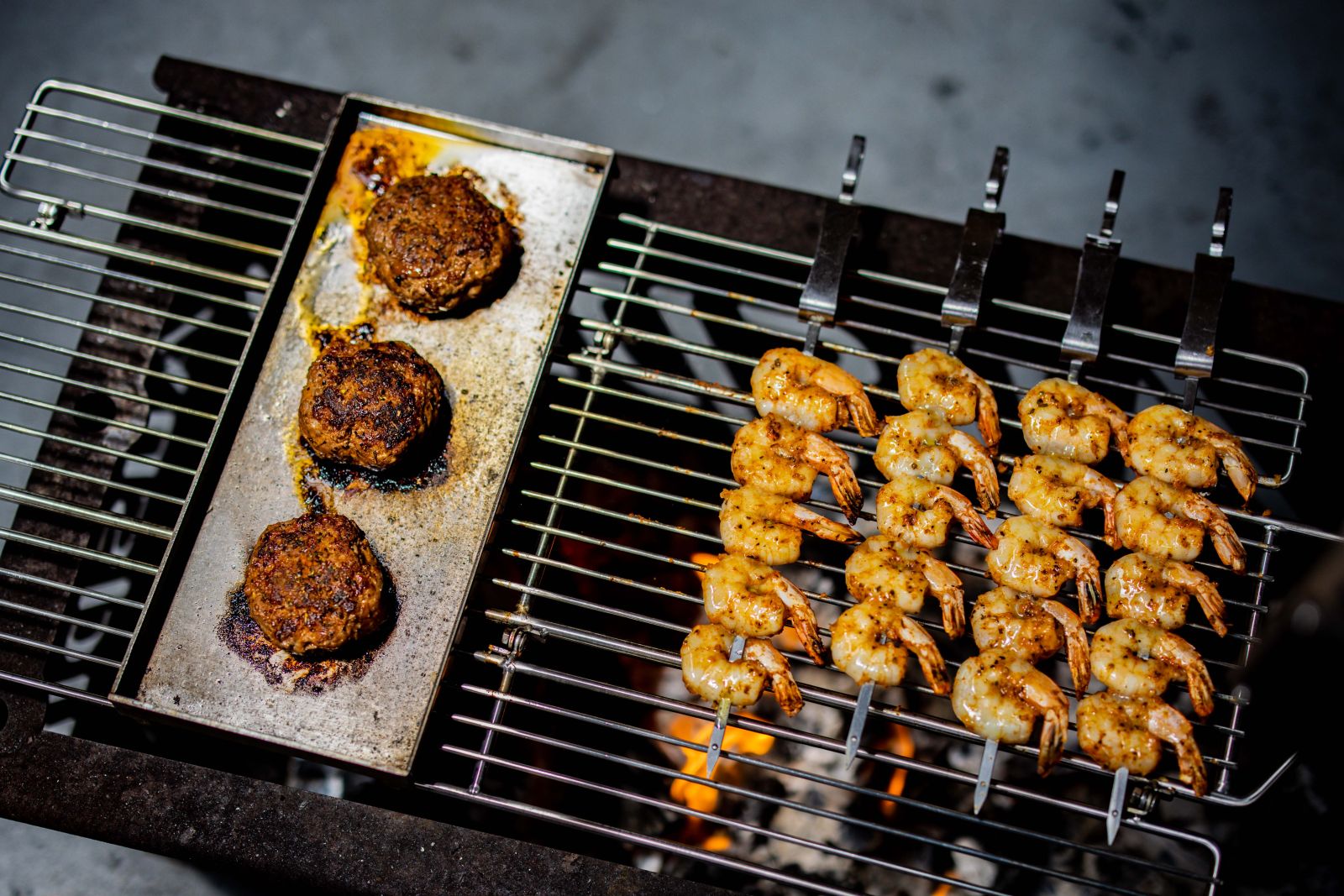 verhead view of prawns and burger patties cooking on an Auspit grill over charcoal. Juicy prawns are skewered and evenly spaced on stainless steel rods, while burger patties sizzle on a hotplate beside them, capturing the perfect outdoor BBQ setup. verhead view of prawns and burger patties cooking on an Auspit grill over charcoal. Juicy prawns are skewered and evenly spaced on stainless steel rods, while burger patties sizzle on a hotplate beside them, capturing the perfect outdoor BBQ setup.