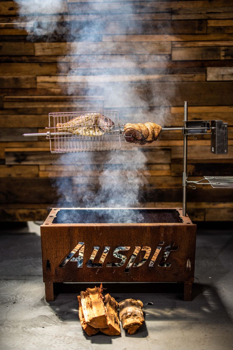 Auspit rotisserie cooking leg of lamb and snapper over charcoal, with smoke rising and wood logs in front.