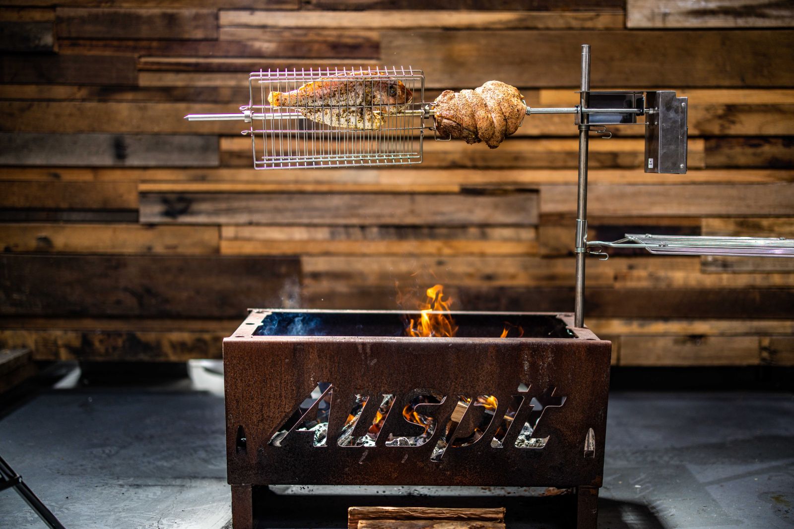 The image shows an Auspit Rotisserie Kit with a leg of lamb and fish on the spit, cooking over a wood-fired base. The rotisserie setup is compact, portable, and ideal for camping or outdoor grilling.