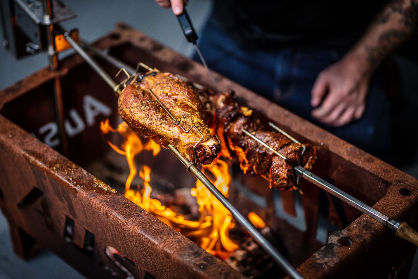 The image shows a close-up of a rotisserie setup with lamb legs roasting over an open flame. The Auspit Rotisserie is positioned above a BBQ grill, with flames visible below, indicating the cooking process in action. The person in the background is holding a thermometer to check the meat's temperature.