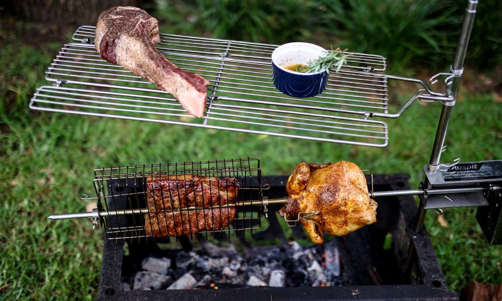 Auspit rotisserie with chicken on prongs, pork roast in a rotisserie basket, and tomahawk steak on the swing-away grill plate above the fire. Auspit rotisserie with chicken on prongs, pork roast in a rotisserie basket, and tomahawk steak on the swing-away grill plate above the fire.