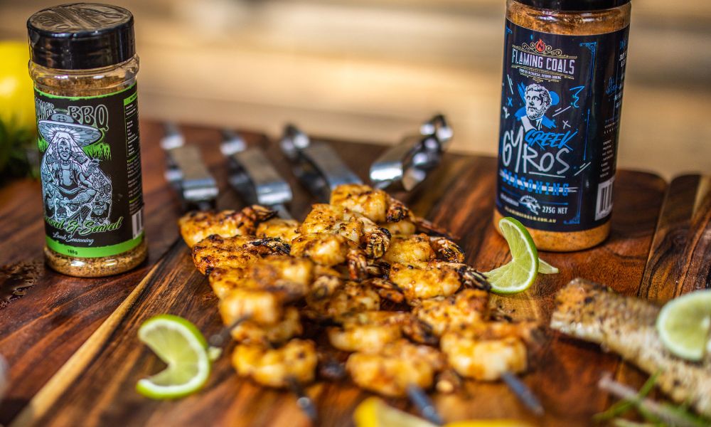 Grilled prawn skewers served on a wooden board with lime wedges, surrounded by Flaming Coals Greek Gyros seasoning and another BBQ rub