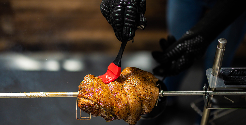 Person wearing heatproof gloves basting a rotating leg of lamb on an Auspit rotisserie with a silicone brush and lemon-olive oil mixture.