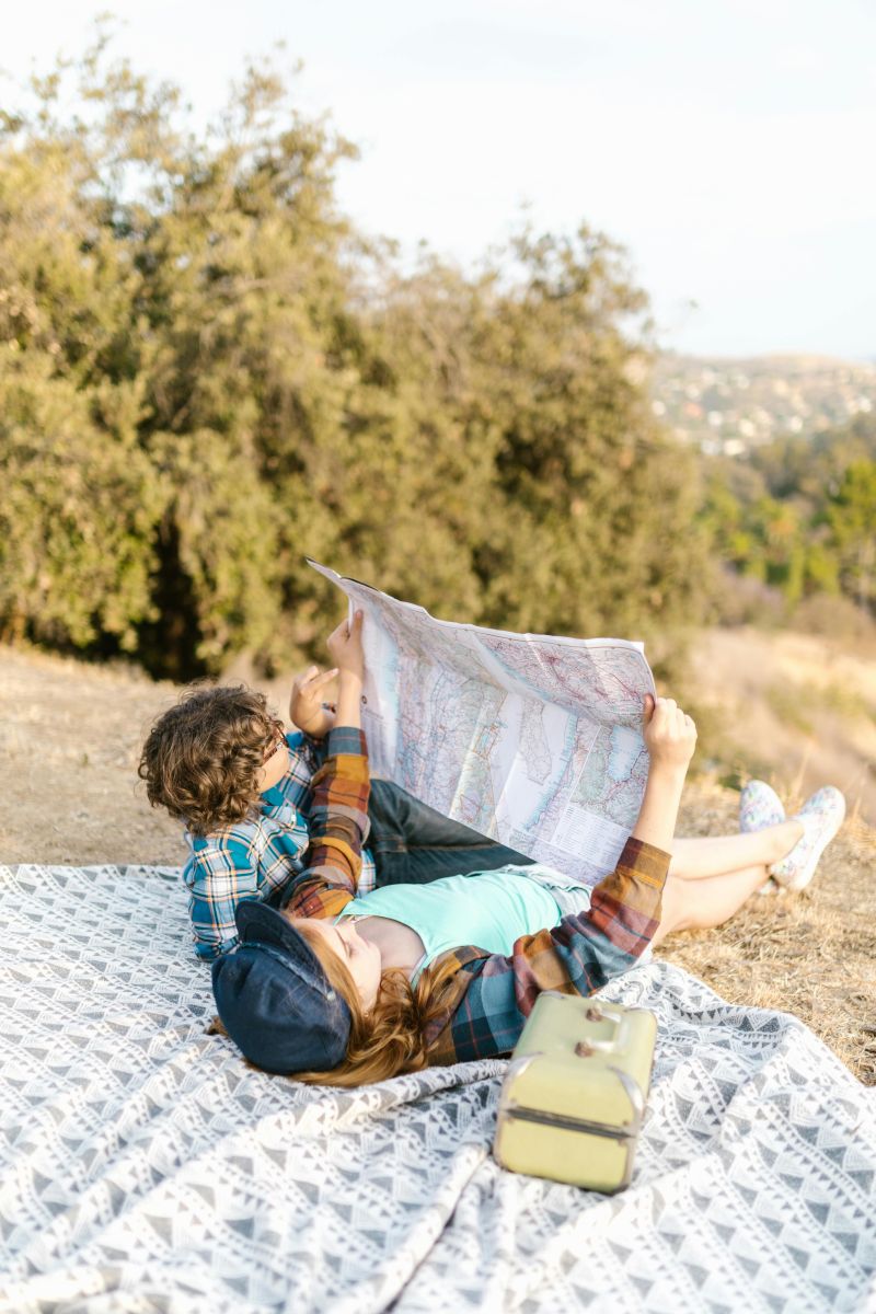 Two kids lie on a picnic blanket outdoors, looking at a large map together. They’re surrounded by dry grass and trees, enjoying a relaxed summer camping moment. Photo by RDNE Stock project: https://www.pexels.com/photo/two-kids-lying-down-on-picnic-blanket-while-looking-at-a-map-8083401/ 