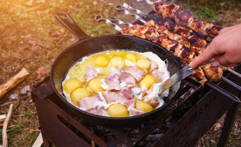 A picture cooking bacon and eggs in a cast iron camp pan over an open fire along side kebabs