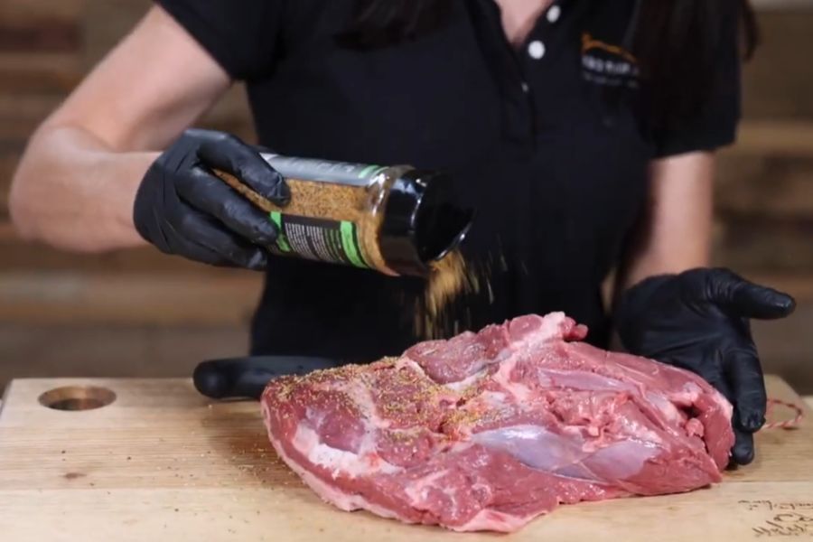 Deboned lamb shoulder being seasoned with Boomas BBQ Sheared & Seared Lamb Seasoning on a wooden board.