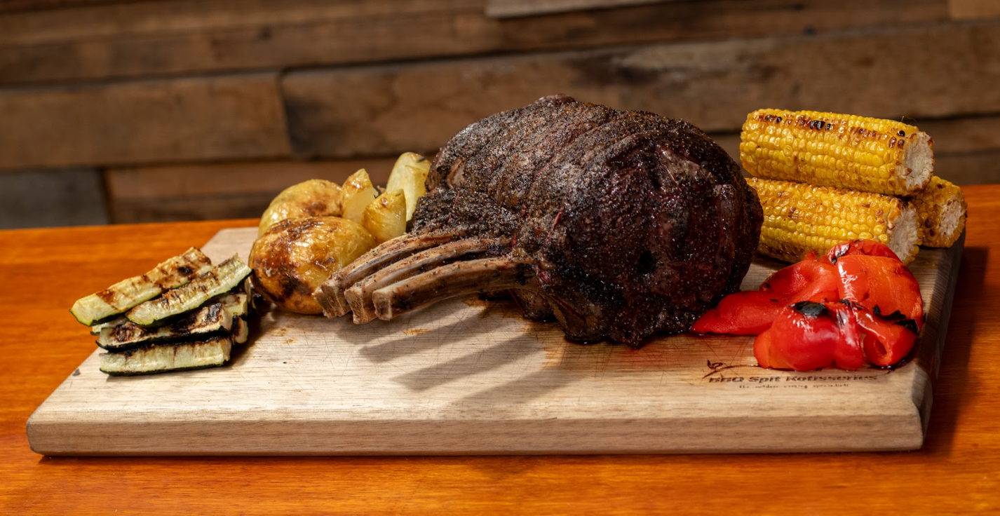Charcoal roasted prime rib served on a wooden board with grilled corn, potatoes, zucchini, and roasted capsicum.