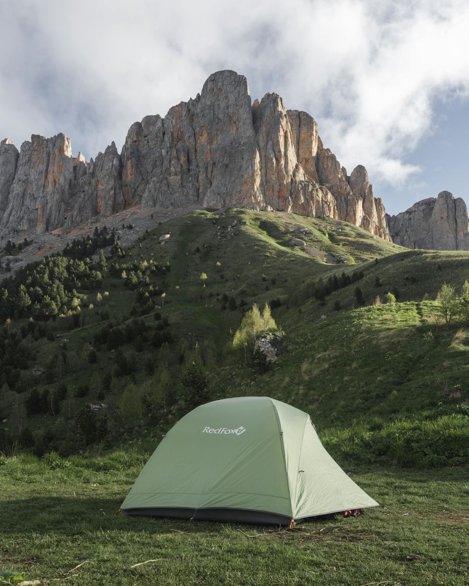 "Green tent set up in a lush meadow, surrounded by towering mountain peaks under a partly cloudy sky, perfect for a spring camping adventure. Source -pexels-greg-betanov