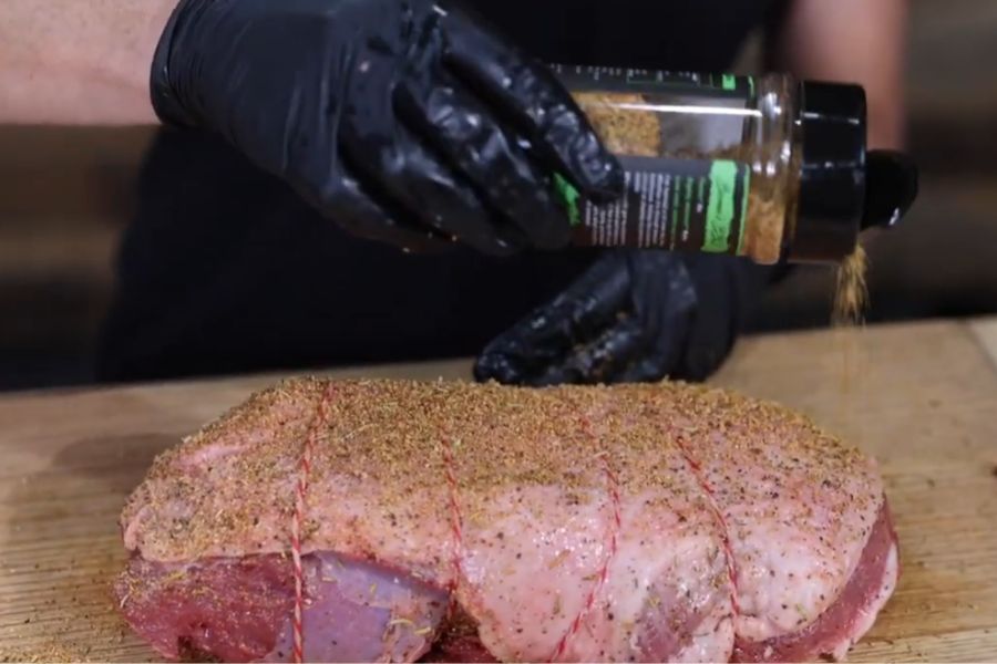Retied deboned leg of lamb being seasoned with Boomas BBQ Sheared & Seared Lamb Seasoning on a wooden chopping board.